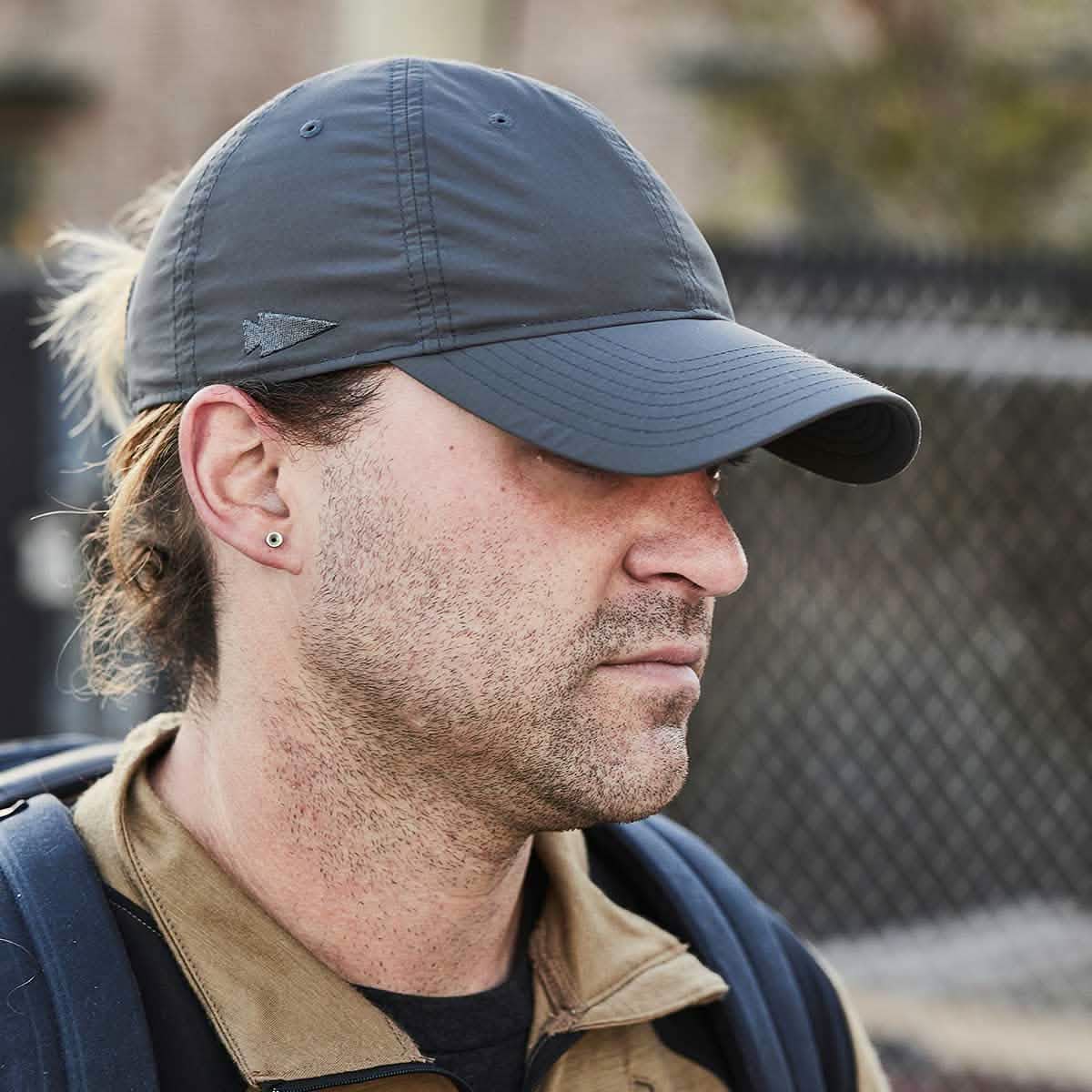 A person wearing a GORUCK Performance TAC Hat - Slick made of TOUGHDRY fabric and a brown jacket is looking away from the camera. They have an earring in the left ear and are carrying a dark backpack. The background shows a blurred fence.