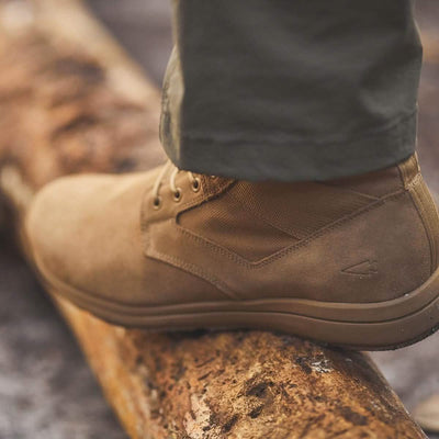 A close-up of the GORUCK MACV-1 - Mid Top in Coyote Suede perched on a weathered log. The person wearing these boots pairs them with olive green pants, highlighting the texture and design against the natural backdrop.
