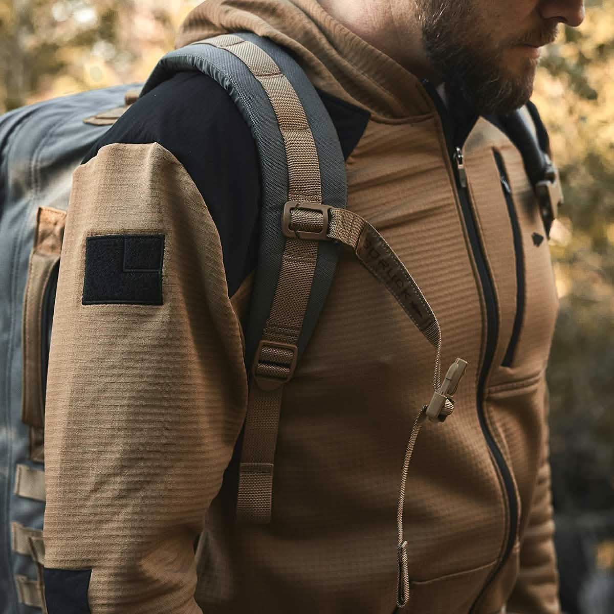 A man in a Men’s Full Zip - Polartec Grid Fleece jacket by GORUCK, featuring fleece lining and ToughDry Fabric for added durability, stands outdoors with a gray backpack. As he explores with the autumn foliage blurred in the background, the backpack straps are visible over his shoulders.