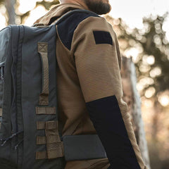 A person wearing the tan and black Men’s Full Zip - Polartec Grid Fleece by GORUCK carries a large green ToughDry Fabric backpack outdoors. The background shows blurred trees, suggesting a natural setting. The focus is on the backpack and the jacket's details.