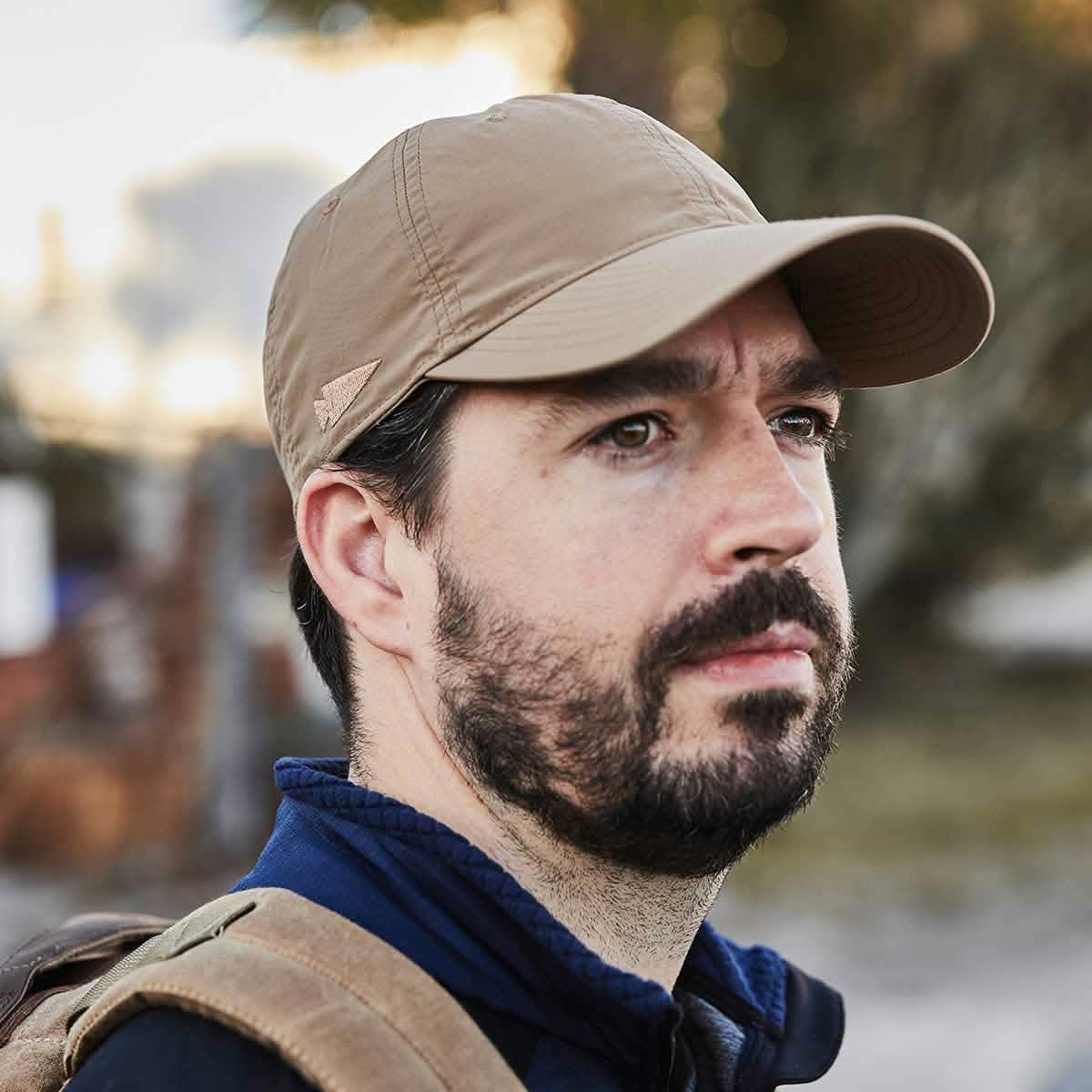 A man with a beard wears the GORUCK Performance TAC Hat - Slick, crafted from TOUGHDRY® fabric renowned for its sweat-wicking abilities. His beige baseball cap beautifully complements his dark jacket as he gazes thoughtfully into the distance. The slight blur in the background suggests an outdoor setting amidst greenery.