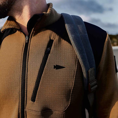 A close-up of a person wearing the GORUCK Men’s Half Zip jacket, featuring a brown design with black accents and a triangle logo, crafted from ToughDry Fabric, while carrying a gray strap backpack. The sky is partly visible in the background.