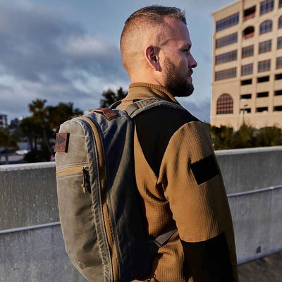 A man with short hair stands outdoors, facing slightly away from the camera. He's wearing a brown jacket crafted from ToughDry Fabric and is carrying a gray backpack. The background showcases a multi-story building beneath a cloudy sky.