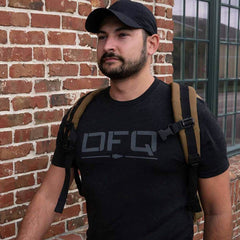 A man with a beard wearing a black cap and a backpack strides confidently past a brick wall with large windows. He is dressed in a DFQ Tee Tri-Blend by vendor-unknown, boldly emblazoned with the message "Don't F*king Quit," capturing his determined spirit.
