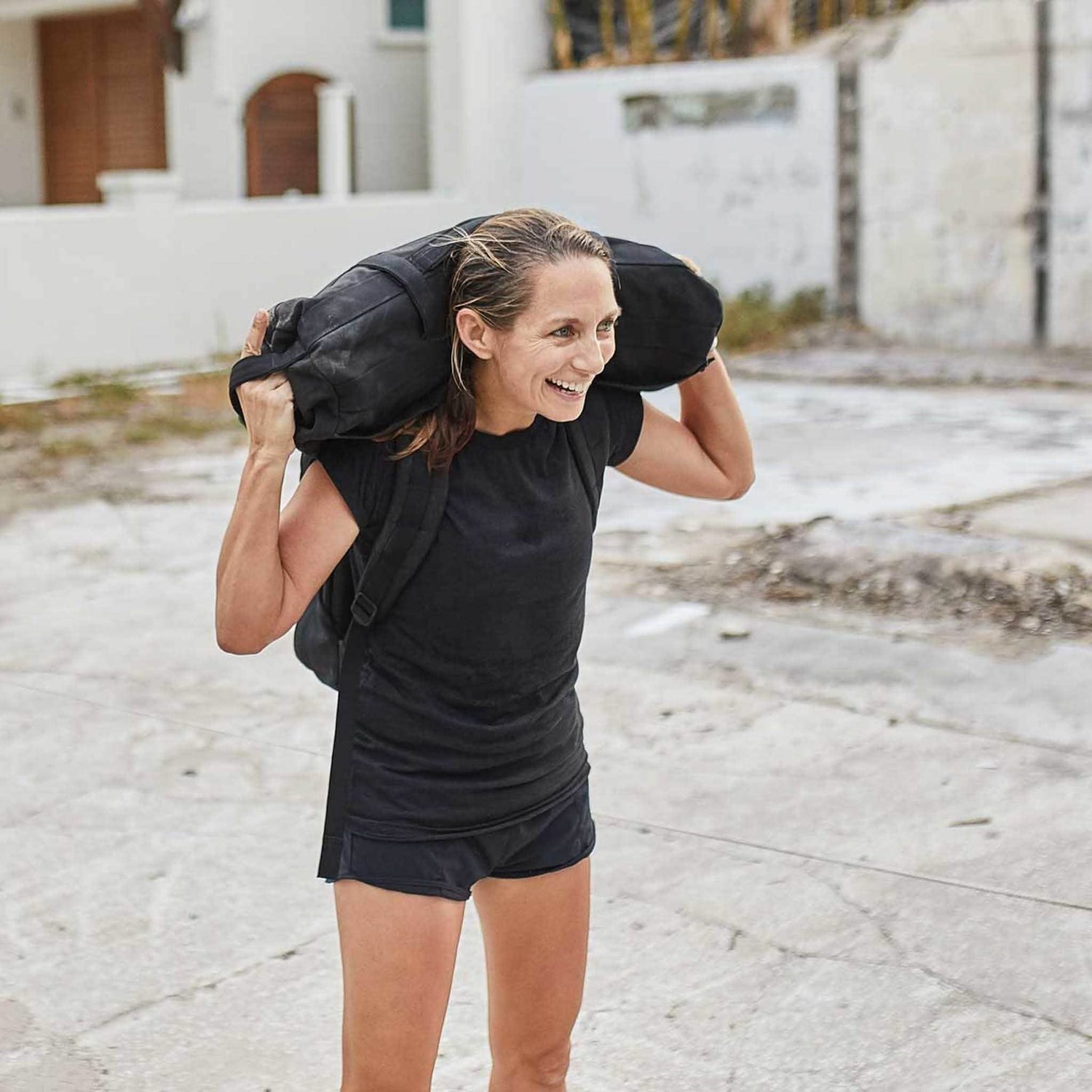 A woman dressed in black athletic wear carries the Sandbags 1.0 on her shoulders outdoors, smiling confidently.