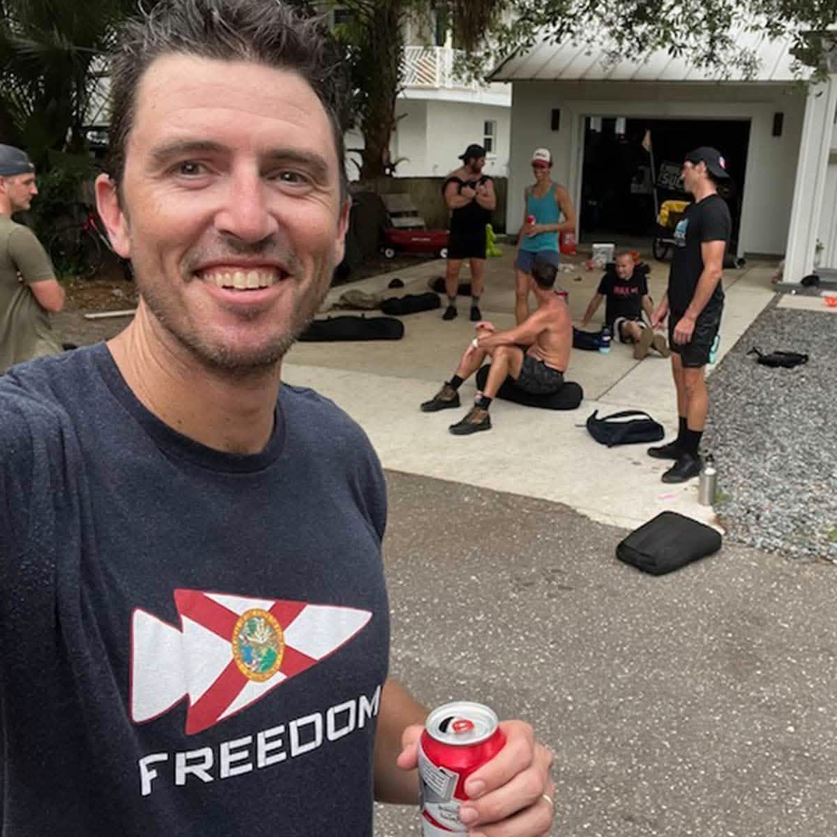 A man beams at the camera, holding a canned drink while wearing a vendor-unknown Freedom Tee - Tri-Blend, proudly displaying the word "FREEDOM." In the backdrop of sunny Florida, a group in athletic gear relaxes and chats on a driveway, with their gear bags resting close by.