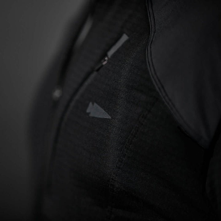 Close-up of a person wearing the Men’s Half Zip - Polartec Grid Fleece by GORUCK, featuring a small, triangular logo on the black fabric. The ToughDry Fabric showcases a textured pattern, and the shirt includes a front zipper. The background is dark gray.