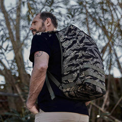 Man wearing a large GR1 - USA - Tiger Stripe rucksack outdoors in front of leafy trees, highlighting its American manufacturing and Special Forces backpack heritage.