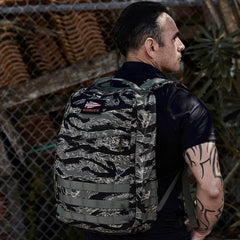 A tattooed man stands by a metal fence outdoors, wearing the GR1 - USA - Tiger Stripe backpack and looking away.