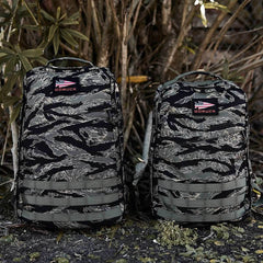 Two GR1 - USA - Tiger Stripe rucksacks stand side by side outdoors, highlighting American manufacturing excellence with lush plants and trees in the background.