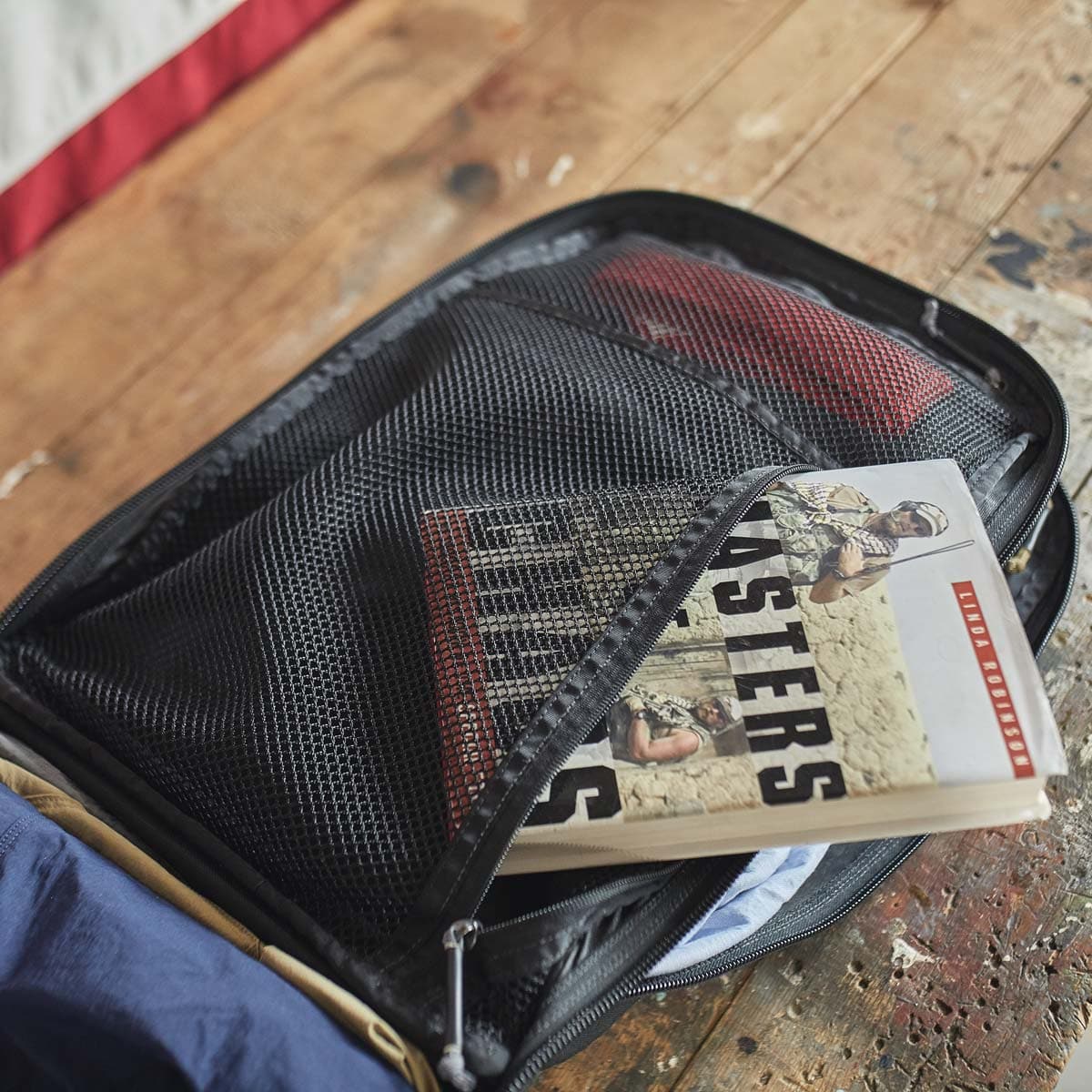 A book partially zipped inside a mesh pocket of an open suitcase on a wooden table.