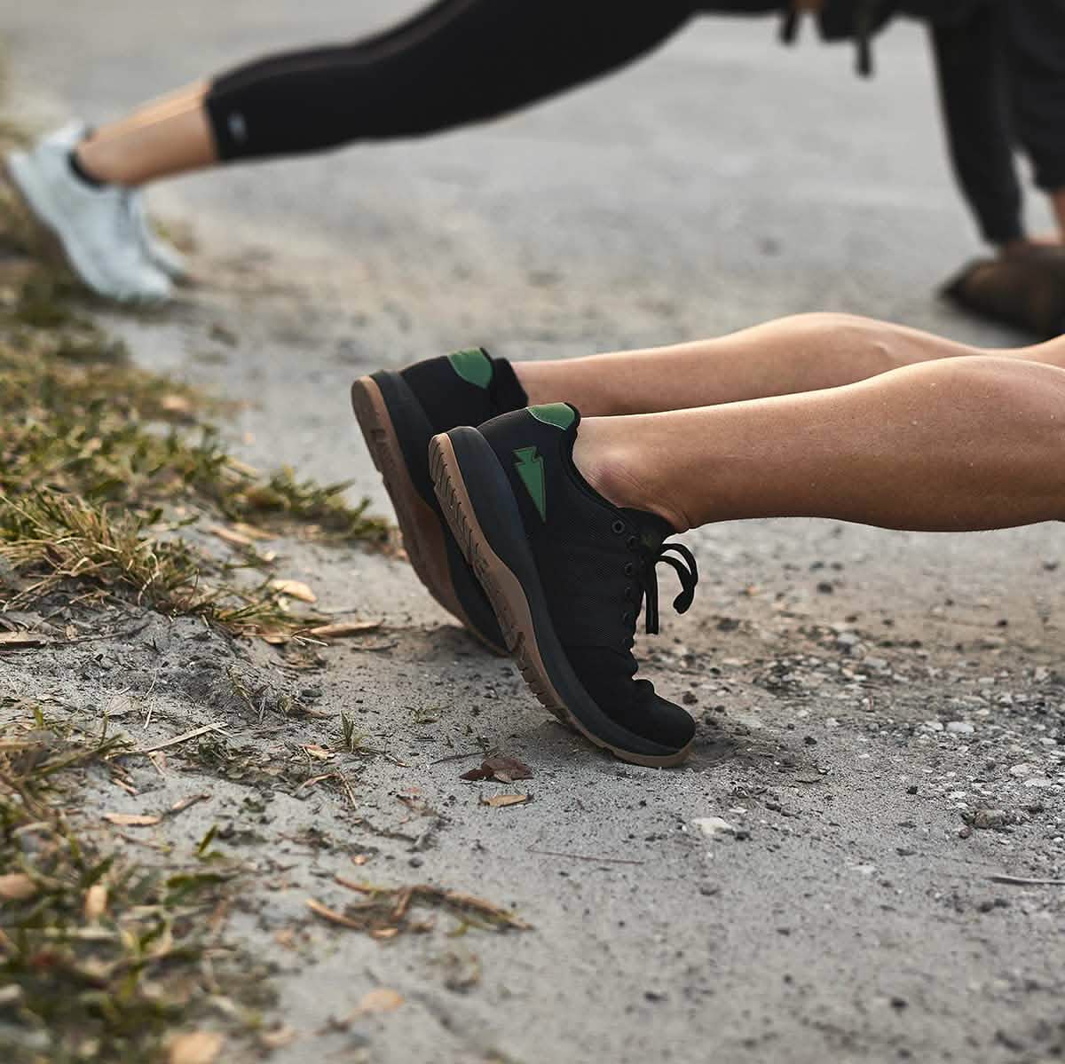 Two people outdoors do push-ups on a paved path, showcasing their athletic legs and Ballistic Trainers - Black + Gum w/ Green Reflective Spearhead—ideal workout shoes for any training session.