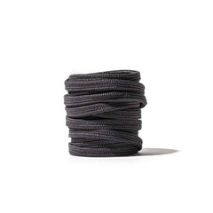 A neatly coiled bundle of GORUCK's Flat Laces in dark gray is positioned against a plain white background, casting a soft shadow on the surface.