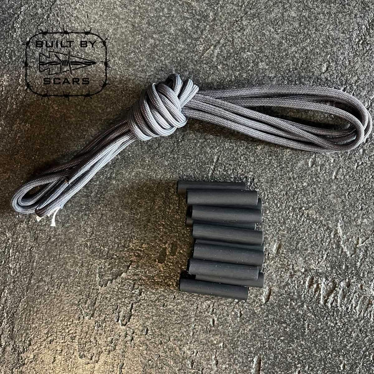 A Paracord Kit - Built By SCARS from GORUCK, featuring a coiled gray paracord and several small black heat shrink tubes ideal for DIY ruck upgrades, is neatly arranged on a textured gray surface with 