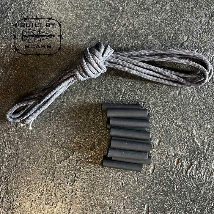 A Paracord Kit - Built By SCARS from GORUCK, featuring a coiled gray paracord and several small black heat shrink tubes ideal for DIY ruck upgrades, is neatly arranged on a textured gray surface with "Built by SCARS" printed in the top left corner alongside the logo.