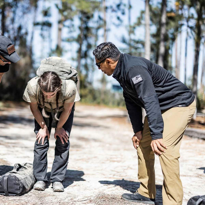Three people stand on a sunlit trail in the forest, with two leaning forward in conversation. The person with a backpack is wearing the GORUCK Men’s Half Zip - Polartec Grid Fleece, making it perfect for outdoor chats. Trees and sky create a serene backdrop.