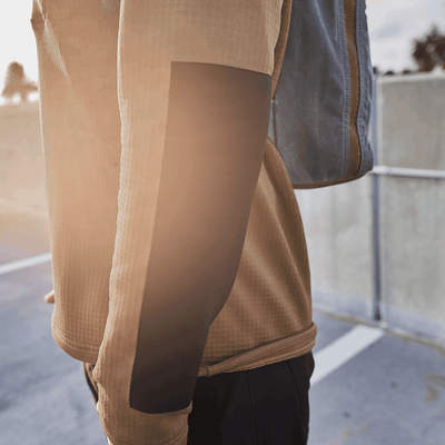 A person wearing the GORUCK Men’s Half Zip in Polartec Grid Fleece walks outdoors, with sunlight casting a warm glow on its sleeve. They carry a gray backpack against a concrete wall, surrounded by soft-focus scenery.