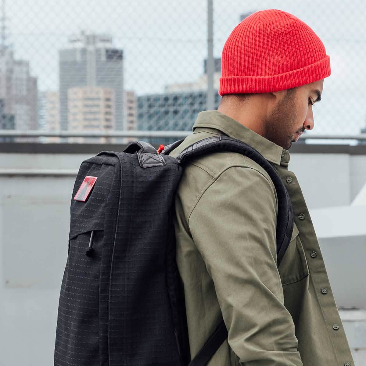 A man in a red beanie and green jacket carries his GR1 USA x Carryology - GRXC2 Samurai backpack through the city.