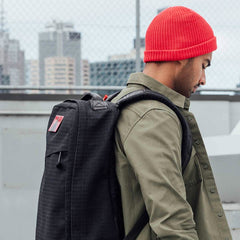 A man in a red beanie and green jacket carries his GR1 USA x Carryology - GRXC2 Samurai backpack through the city.