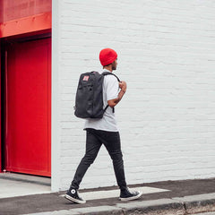 A person in a red beanie and backpack, possibly the GR1 USA x Carryology - GRXC2 Samurai, walks by a white brick wall with a red door.