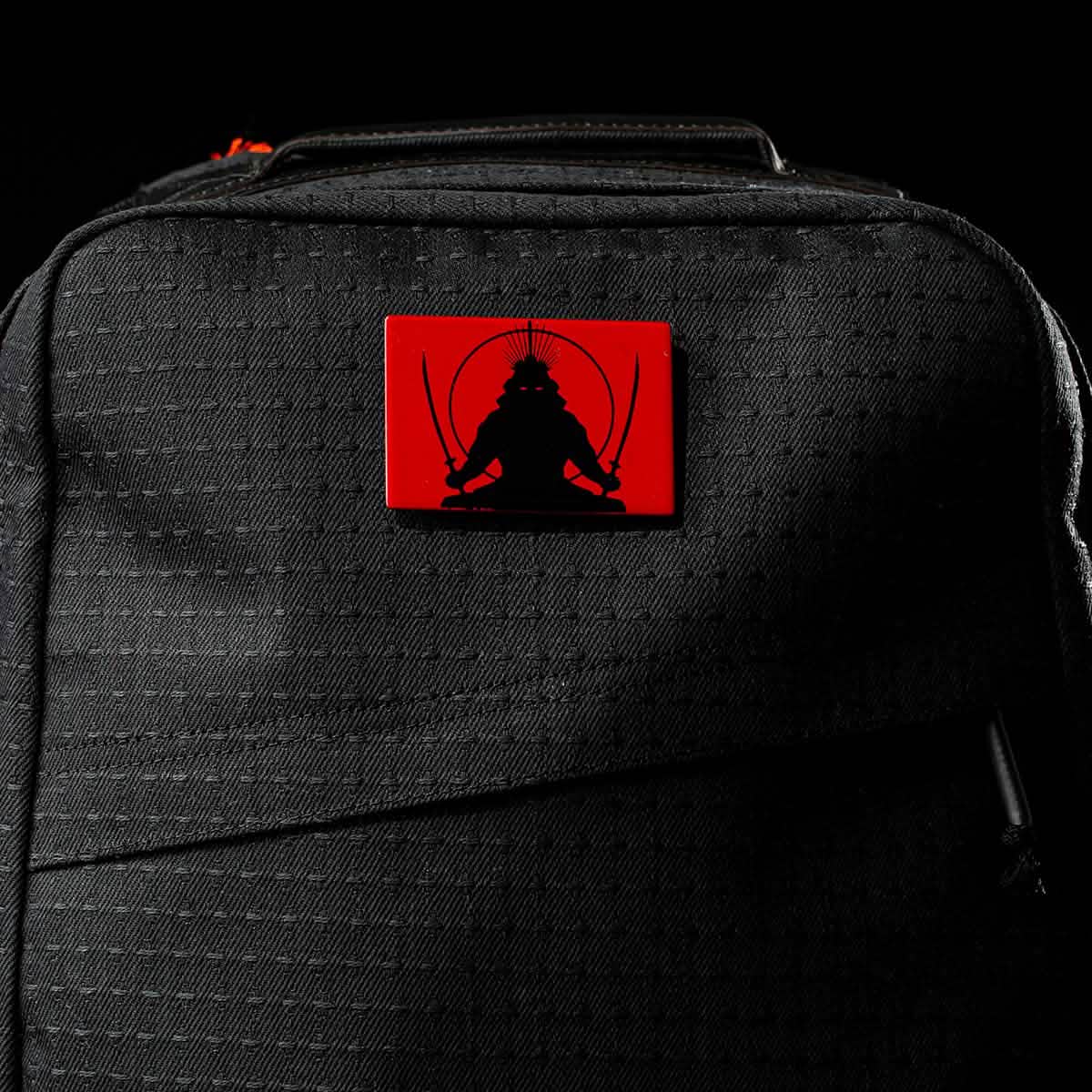 The GR1 USA x Carryology - GRXC2 Samurai is a black backpack with a red patch showing a black samurai silhouette holding two swords, crafted from premium Japanese cotton fabric.