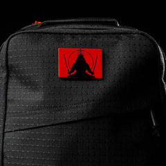 The GR1 USA x Carryology - GRXC2 Samurai is a black backpack with a red patch showing a black samurai silhouette holding two swords, crafted from premium Japanese cotton fabric.