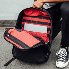 A person opens a GR1 USA x Carryology - GRXC2 Samurai backpack with a bold red interior, revealing a laptop inside, while wearing black sneakers.