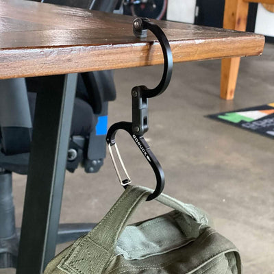 A black Heroclip - GORUCK Logo gear clip is attached to the edge of a wooden table, securely holding a green bag. Designed by Heroclip with 360° versatile rotation, this hook is perfect for hanging bags to keep them off the floor. In the background, you can see a chair and part of a mat.