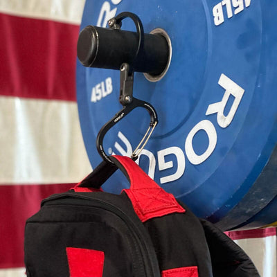 A black and red backpack is hanging from a Heroclip - GORUCK Logo attached to a 45LB Rogue weightlifting plate. Featuring versatile 360° rotation, the Heroclip holds fast as an American flag waves proudly in the background.