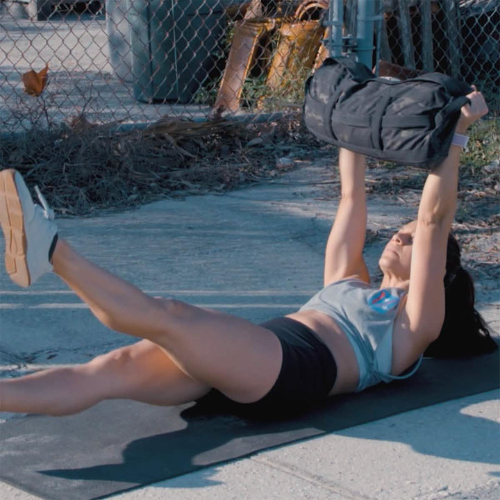 A person outdoors is performing a leg raise while lying on their back, holding a sandbag from the GORUCK brand's "The Pushover with Jaala Shaw (4-Week Training Program)" above their chest. One leg is extended upward, and the other slightly off the ground. This exercise is ideal for consistent strength building, set against a background of a chain-link fence and vegetation.