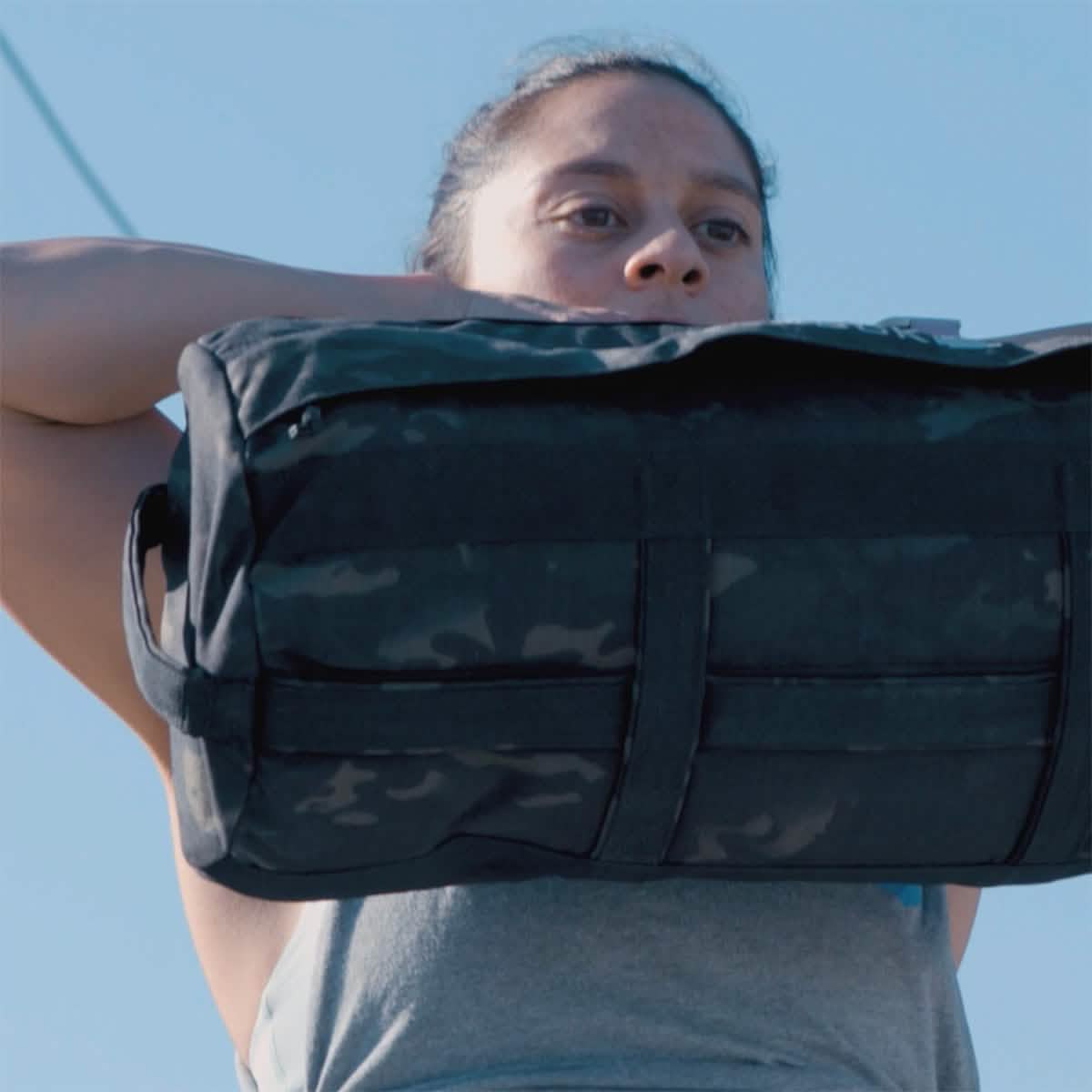 Demonstrating the results of The Pushover with Jaala Shaw, a 4-week training program by GORUCK, a focused and determined individual effortlessly lifts a large black duffel bag over their shoulders outdoors. Clad in a gray sleeveless top against the backdrop of a bright blue sky, they showcase their consistent strength building.