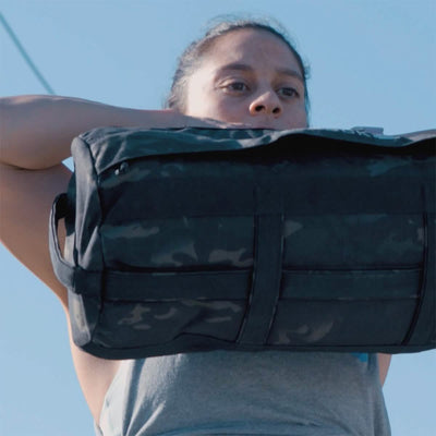 Demonstrating the results of The Pushover with Jaala Shaw, a 4-week training program by GORUCK, a focused and determined individual effortlessly lifts a large black duffel bag over their shoulders outdoors. Clad in a gray sleeveless top against the backdrop of a bright blue sky, they showcase their consistent strength building.