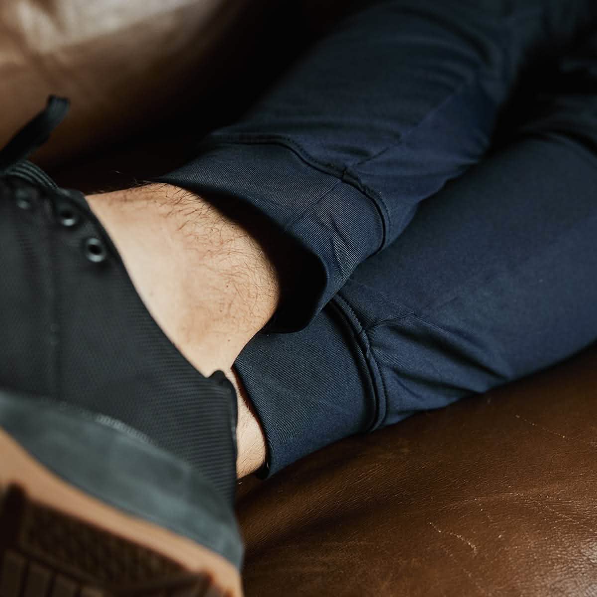 Close-up of a person wearing dark blue GORUCK Indestructible Performance Joggers and black sneakers, resting on a brown leather surface. The image focuses on their crossed legs, highlighting the moisture-wicking performance fabric's texture and the color of the clothing and footwear.