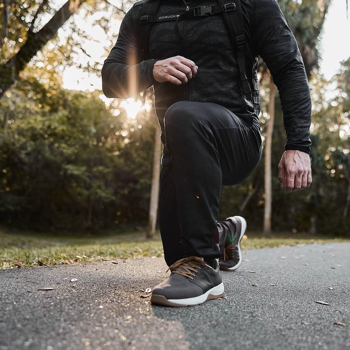A person in athletic gear is kneeling on a paved path in a park, with a backpack on. Sunlight filters through the trees, highlighting their laced Indestructible Performance Joggers by GORUCK, made from performance fabric that promises comfort and comes with Scars Lifetime Guarantee.