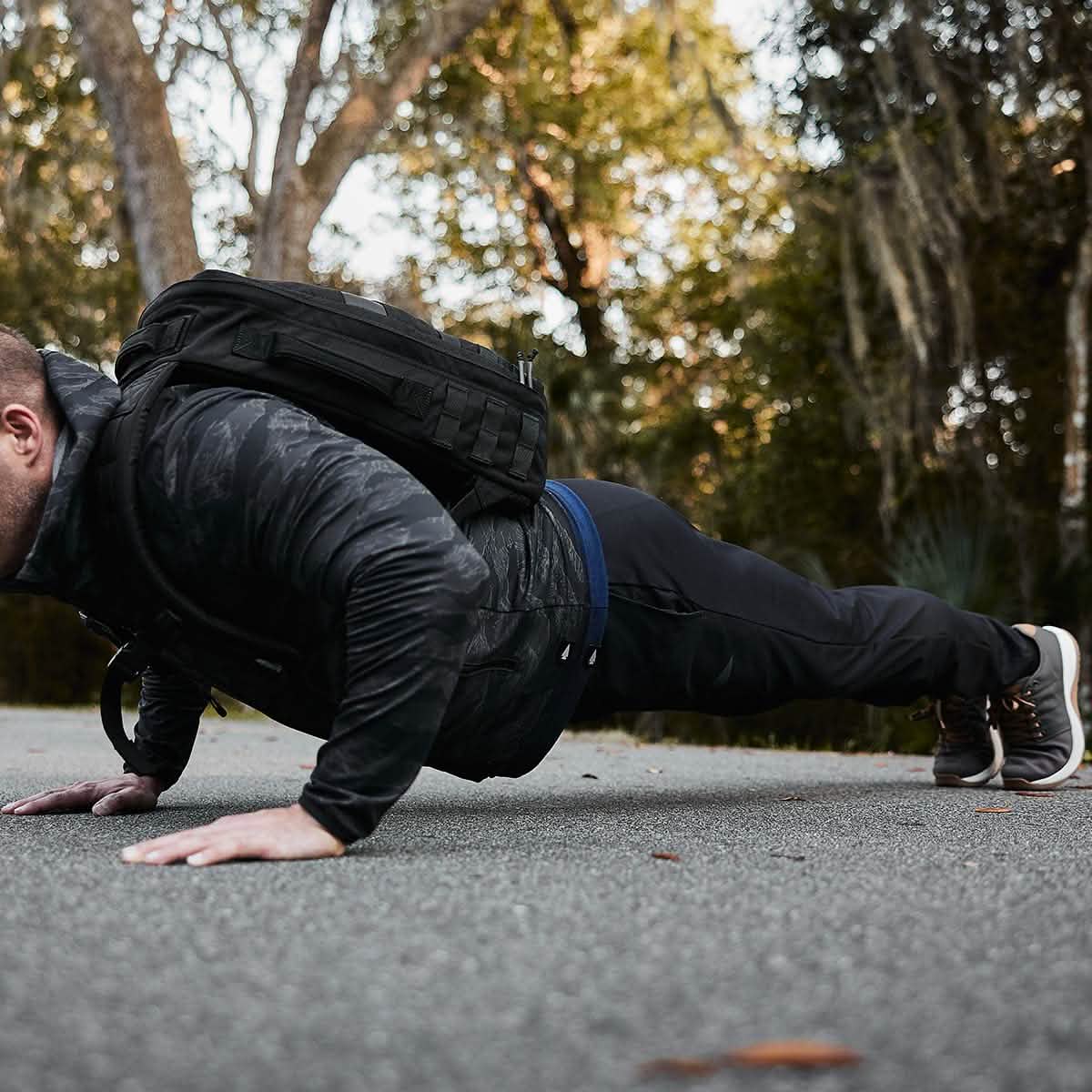 An individual in athletic attire demonstrates their strength on a paved path through the woods. Wearing a camo jacket, GORUCK Indestructible Performance Joggers, sneakers, and a black backpack crafted from performance fabric, they seamlessly blend into the lush green surroundings.
