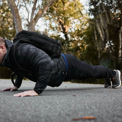 An individual in athletic attire demonstrates their strength on a paved path through the woods. Wearing a camo jacket, GORUCK Indestructible Performance Joggers, sneakers, and a black backpack crafted from performance fabric, they seamlessly blend into the lush green surroundings.