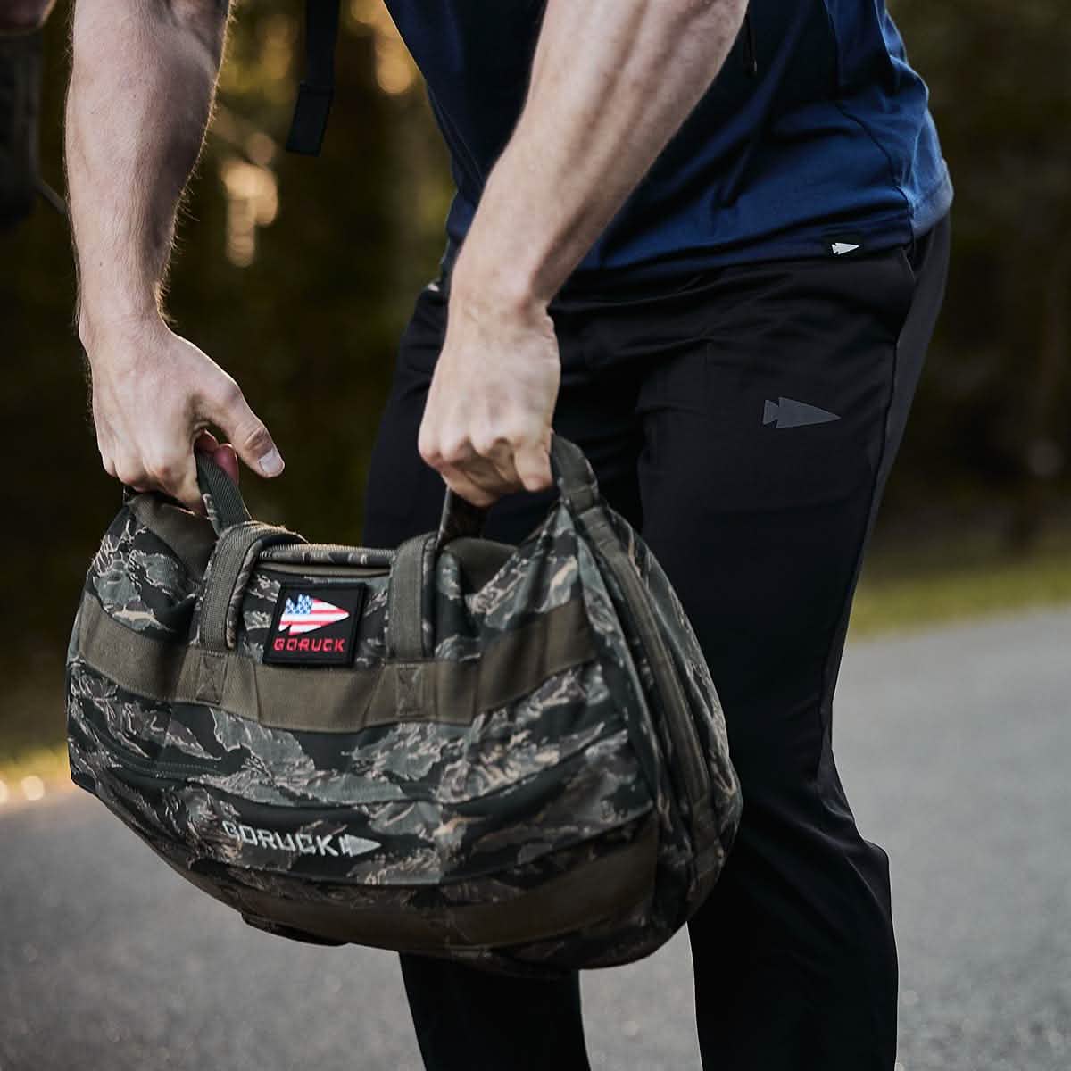 A person dressed in GORUCK Indestructible Performance Joggers lifts a camouflage-patterned GORUCK bag with both hands outdoors. Made from performance fabric and featuring a patch with the American flag, this durable bag is built to last and comes with the Scars Lifetime Guarantee. The background consists of blurred greenery.