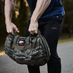 A person dressed in GORUCK Indestructible Performance Joggers lifts a camouflage-patterned GORUCK bag with both hands outdoors. Made from performance fabric and featuring a patch with the American flag, this durable bag is built to last and comes with the Scars Lifetime Guarantee. The background consists of blurred greenery.