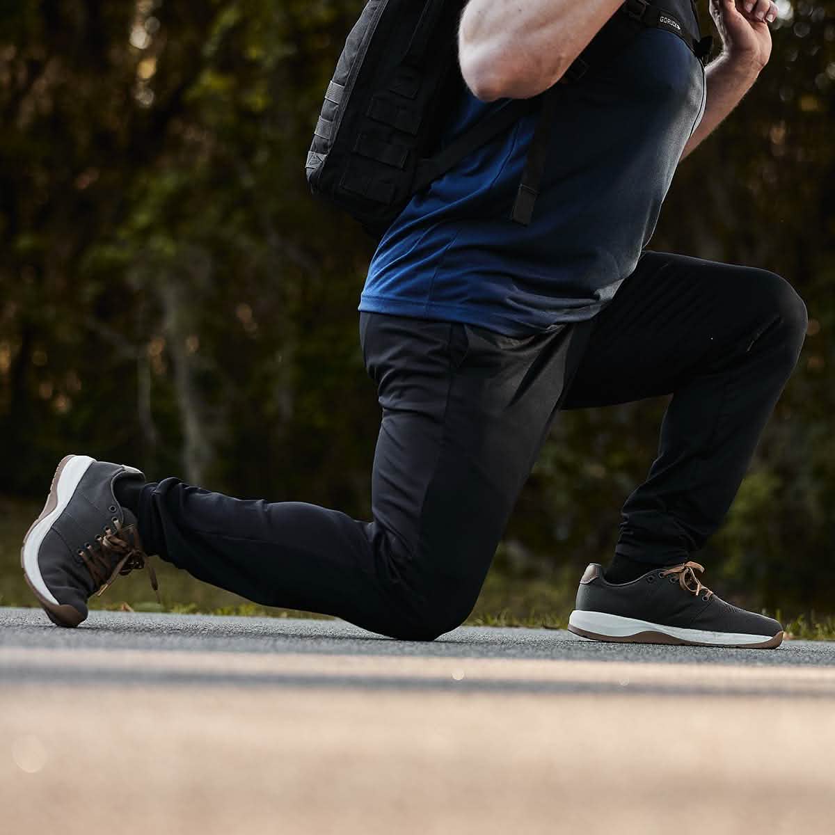 A person dressed in athletic attire and carrying a backpack is kneeling on one knee on a road. Wearing GORUCK's Indestructible Performance Joggers crafted from high-performance fabric, the emphasis is on the footwear and posture, set against a blurred greenery background.