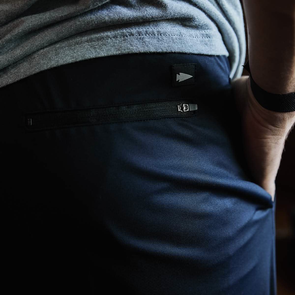 A close-up of a person wearing GORUCK Indestructible Performance Joggers in navy blue, featuring a small logo on the pocket. The individual is also sporting a gray t-shirt and a black wristband while standing against a dark background.