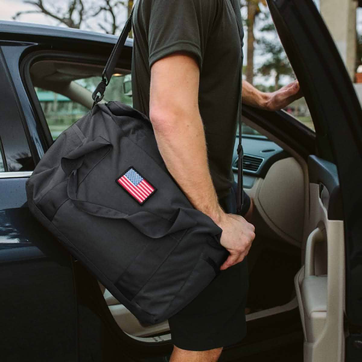 A person with a 32L Kit Bag - USA, displaying a US flag patch and proudly made in the USA, stands by an open car door.