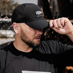 Man wearing a black GORUCK trucker hat with logo patch, outdoor urban background