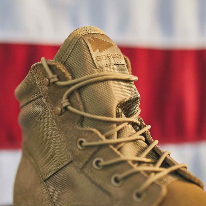 A close-up shot of a MACV-1 - Mid Top boot in coyote suede showcases its military style, crafted with a wide toe box. The brand name GORUCK is prominently displayed on the tongue, while the all-terrain outsole stands out against a background blurred with red and white stripes.