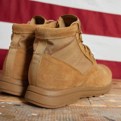 A pair of MACV-1 - Mid Top boots in coyote suede from GORUCK are displayed on a wooden surface, highlighting their durable material and rugged design. The all-terrain outsole suggests excellent durability, while in the background, a blurred red and white striped pattern provides a vibrant contrast.