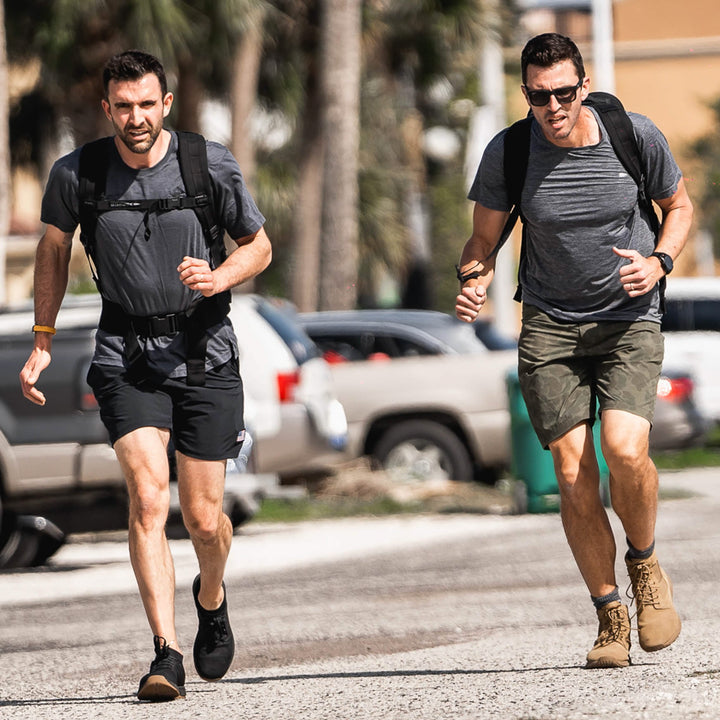 Two men are jogging on a sunlit street, each sporting backpacks reminiscent of the vendor-unknown brand and casual athletic wear. One wears black shorts while the other opts for green. Their fitness goals propel them past parked cars and palm trees in the background.