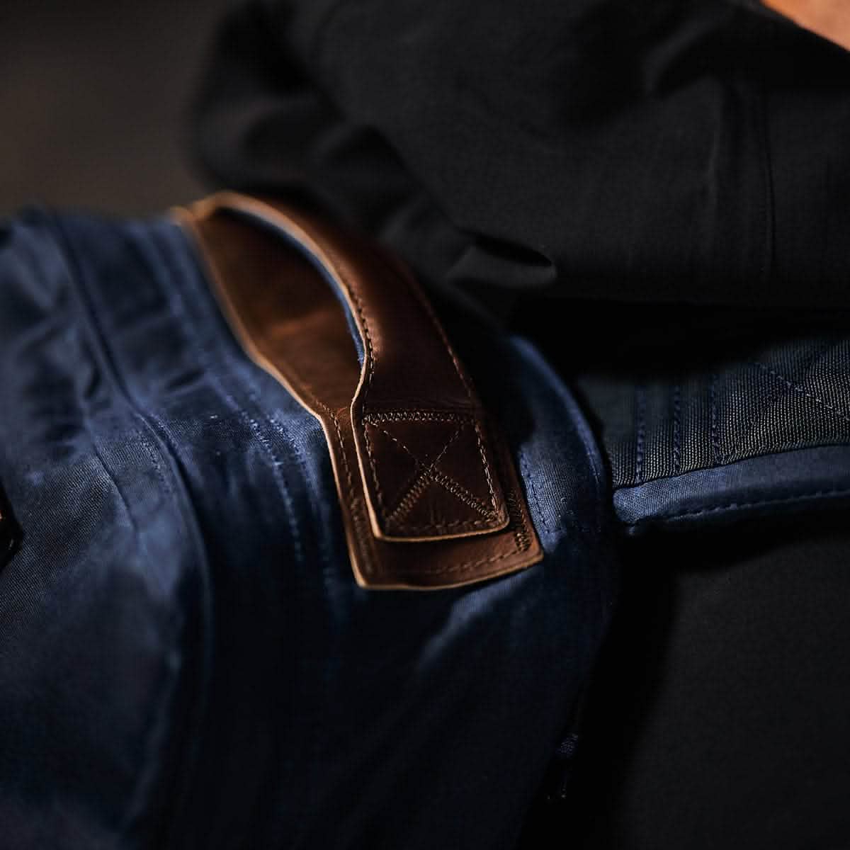 Close-up of a person wearing a dark blue GR1 USA - Heritage rucksack by GORUCK with a brown Red Wing Leather handle on their shoulder. The bag and handle are contrasted against a black jacket, highlighting the textures and materials.