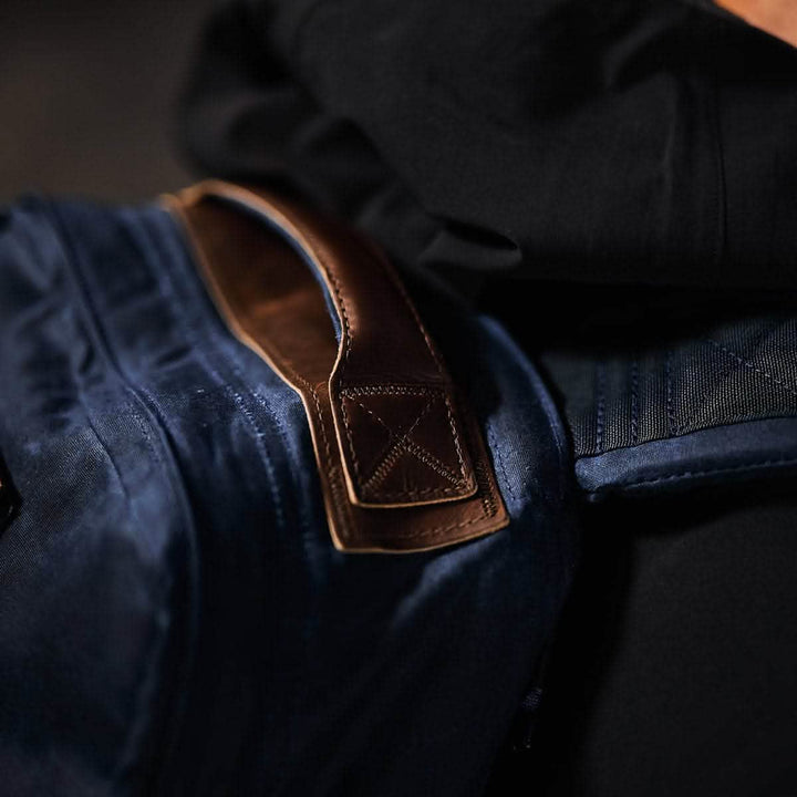 Close-up of a person wearing a dark blue GR1 USA - Heritage rucksack by GORUCK with a brown Red Wing Leather handle on their shoulder. The bag and handle are contrasted against a black jacket, highlighting the textures and materials.