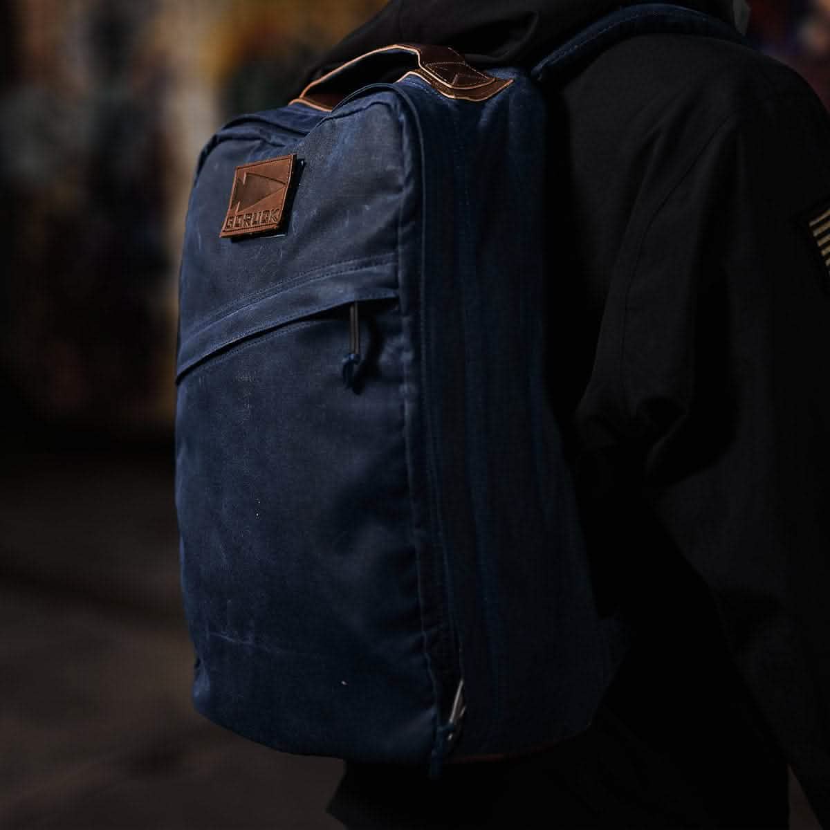 A person wearing a dark jacket and carrying a GORUCK GR1 USA - Heritage, crafted from Waxed Duck Canvas, stands in a dimly lit environment. The soft lighting sets a moody atmosphere.