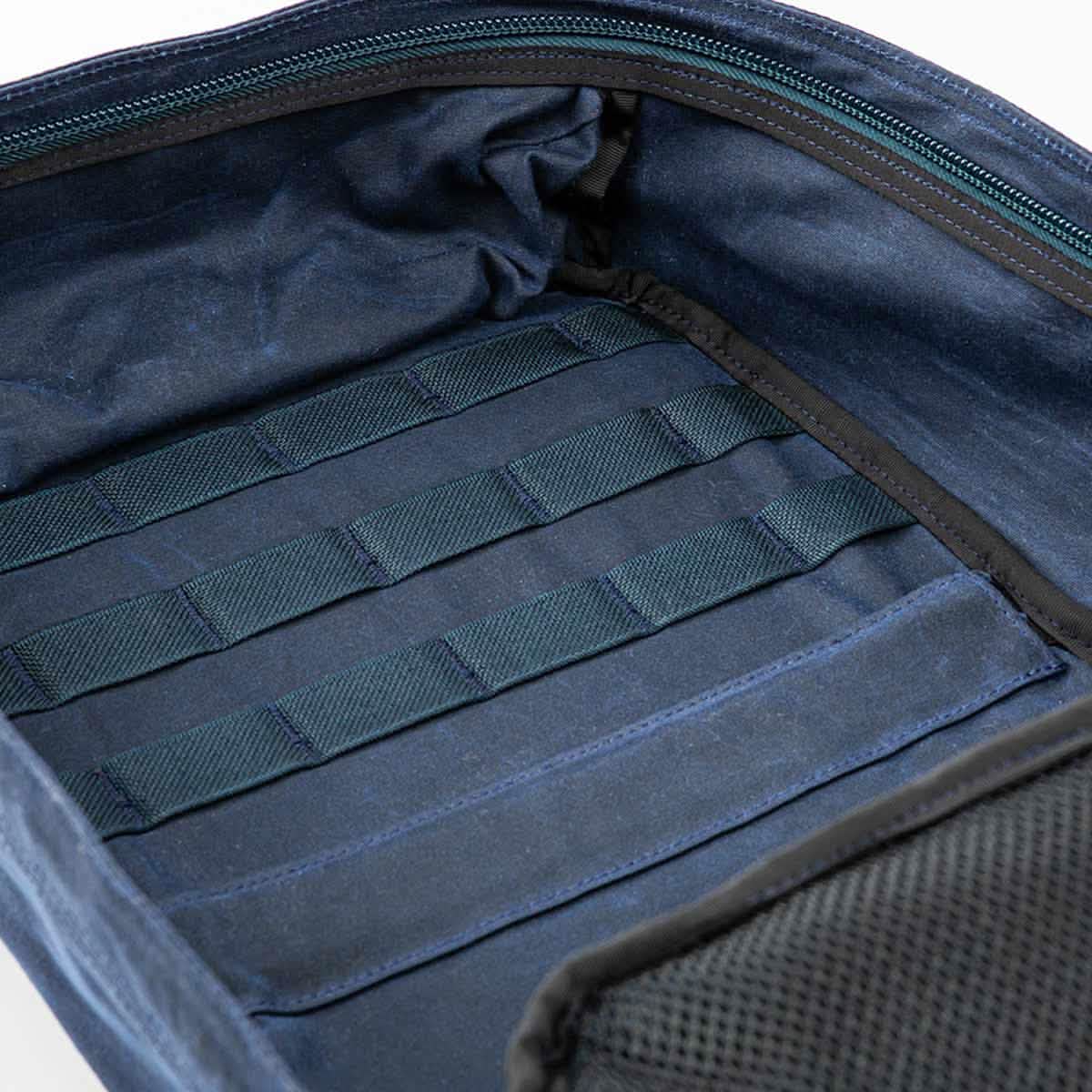 The GR1 USA - Heritage from GORUCK showcases a waxed duck canvas exterior, which opens to disclose a blue fabric interior featuring greenish horizontal straps for organizing your belongings. Inside, the mesh-front pocket adds both convenience and style, ensuring that this product remains functional and timeless.