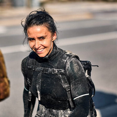 A person wearing a GORUCK jacket adorned with MOLLE webbing beams while covered in mud on a street. They have a backpack and appear to be participating in a rugged outdoor activity, using their Training Sternum Strap. The background features a blurred road.